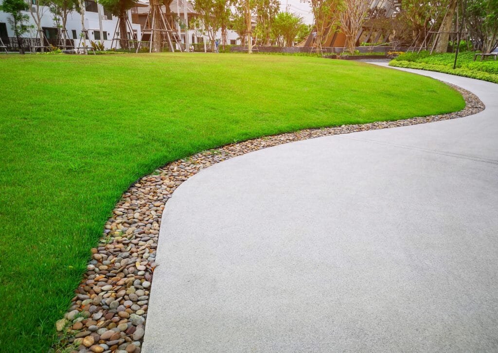 Smooth green grass lawn with gray curve pattern walkway, sand-washed finishing on concrete paving with brown gravel border, trees with supporting and shrubbery - turf installation by Zebrascapes