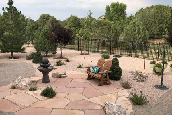 An outdoor living space with a combination of pavers, natural stone hardscape, and pebbles. Native plants and shrubs are scattered throughout and there is a bench in the middle. You can see a pergola and a fire pit in the distance